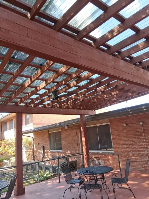 A stained wood pergola with a corrugated polycarbonate roof and ceiling fan, built against a brick home.
